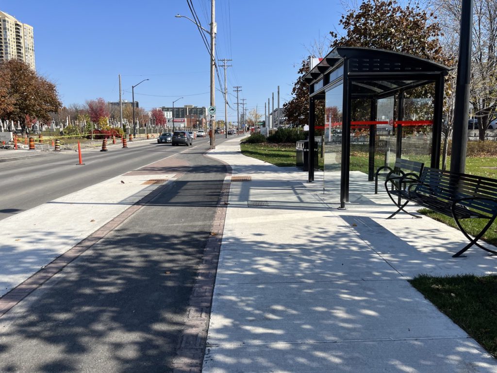 Picture of bicycle pathway running between the sidewalk and a place for people waiting for the bus to queue.