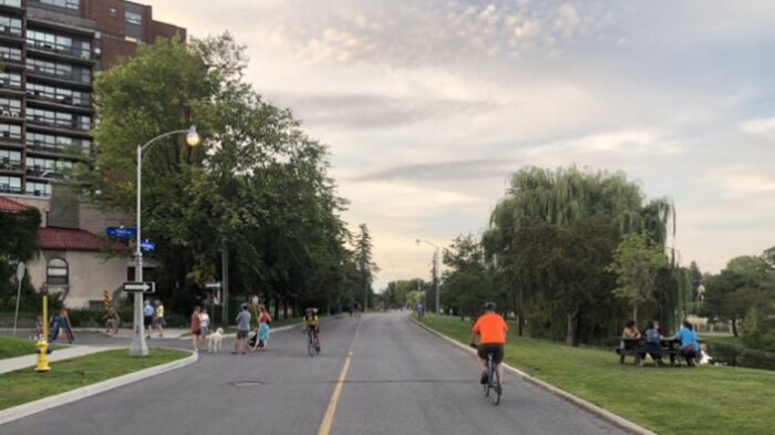 View along a street of a group of people with a dog standing in the roadway talking with cyclists passing in both directions. On the opposite side of the street, people are sitting at a picnic table next to a pond with trees. The photo is taken near sunset and there are thin clouds in the sky.