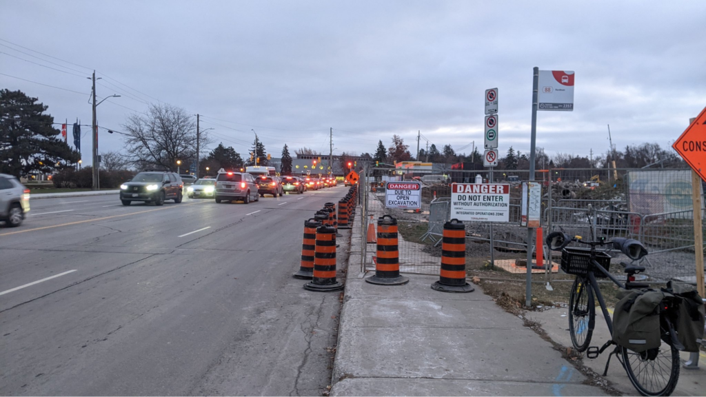 A sidewalk blocked by construction fencing, with a line of traffic cones arranged along the curb.