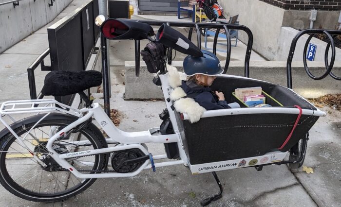 Cargo bike parked alongside coat hanger style of bike rack (which doesn’t fit cargo bikes). There’s a child sitting in the bin at the front.