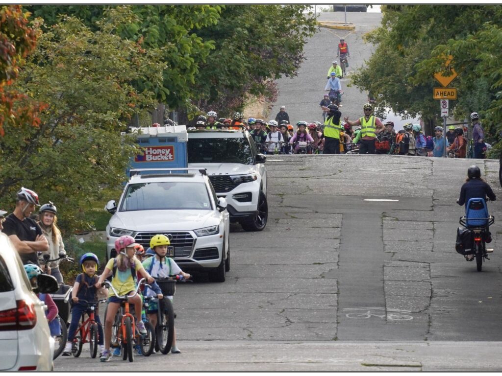 Image of a bike bus over rolling hills. In the background, kids and adults rolling down the hill. In the foreground, on the left, a group of kids waiting for the bike bus to arrive to join.