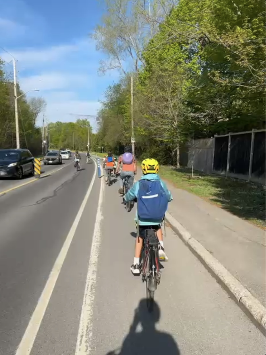 Les parents et enfants du vélo bus circulent en file dans la voie cyclable de la rue Hemlock. Deux personnes à vélo au loin les dépassent et à gauche on peut voir des voitures qui circulent dans l’autre sens. Quelques piquets flexibles séparent la voie cyclable du couloir des voitures. Un large piquet sépare les deux couloirs de voitures.