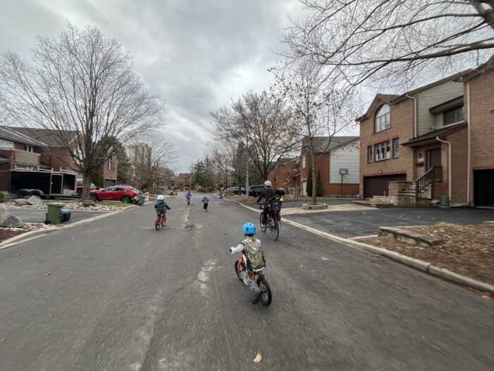 One adult and four children rolling down a quiet neighbourhood street, one of them on a scooter. Trees line up the street on both sides