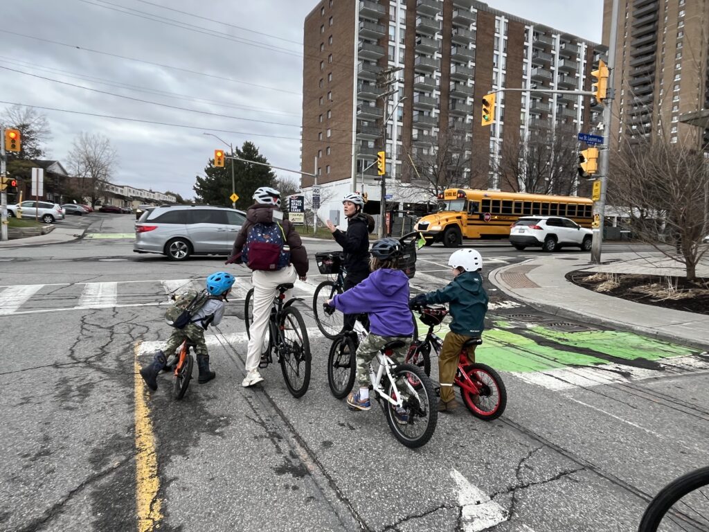 Two adults and three kids waiting at a stop line in a left turning lane for green light. Traffic moving in the background on Saint-Laurent.