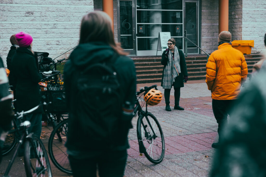 Councillor Ariel Troster addressing the Bill 60 rally. People and their bikes can be seen in the foreground and to the left.