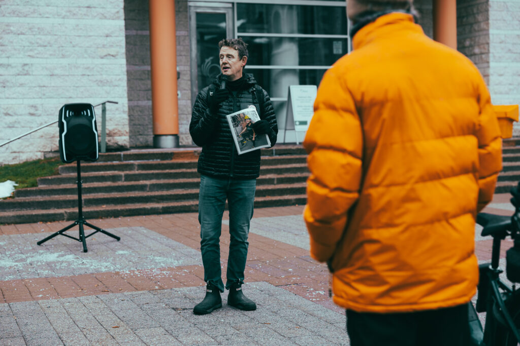 Former MPP Joel Harden addressing the Bill 60 rally. A person is listening in the foreground.