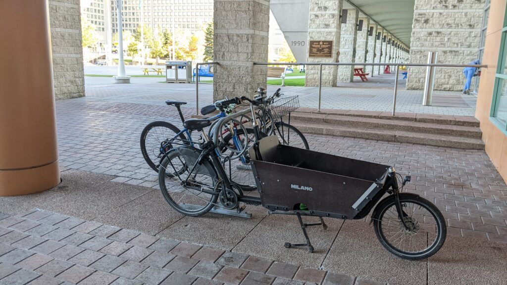 Picture of several bicycles at city hall, several standard bicycles are at the bicycle rack, however one cargo bike is locked to the outside of the rack, due to the rack not being able to accommodate the size of the bicycle.
