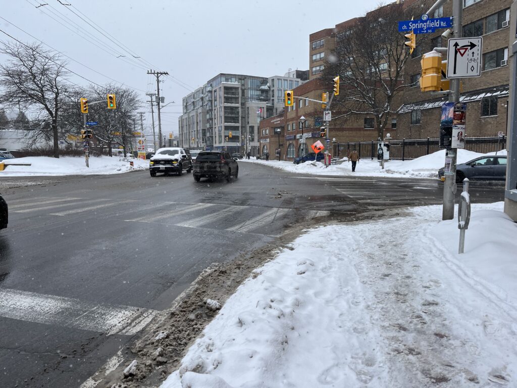 Picture of an intersection with no right-turn-on-red restriction and a yield-to-cyclist sign for drivers turning right. Painted bike lane is snowed in and not visible